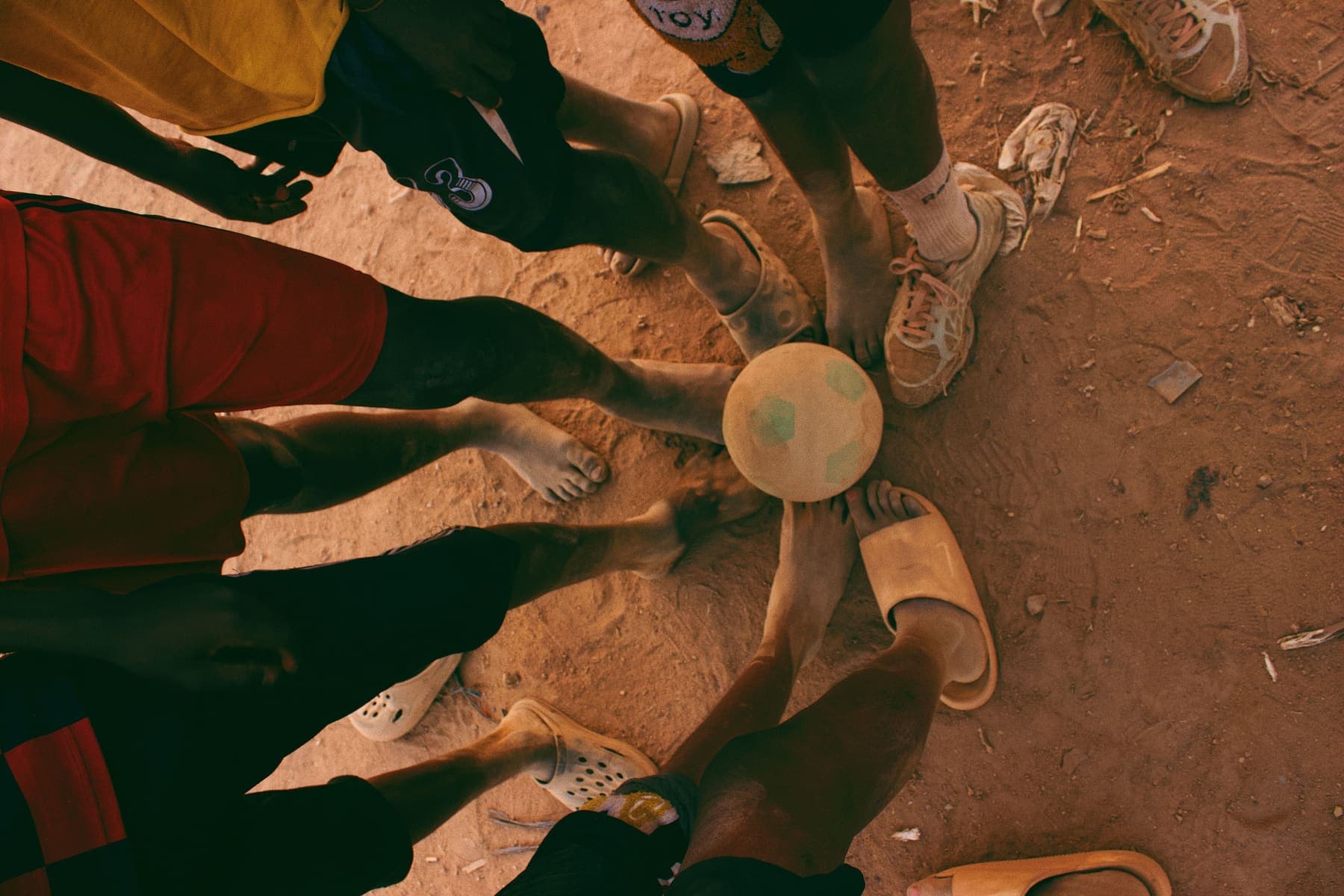 Kids gathered in a circle around a football, feet touching the ball, playing barefoot on dirt ground